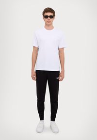 Witte kortegemouwen t-shirt, zwarte joggingsbroek met een logo, en witte sneakers; model draagt zwarte zonnebril, staat voor een neutrale achtergrond.
