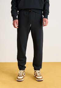 Sorte sweatpants lavet af blødt stof, med en elastisk talje med snøre og bukseafslutninger, parret med brune og cremefarvede sneakers.
