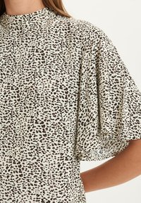 Blus med leopardmönster, hög hals och korta, fladdrande ärmar. Tyget har en krämfärgad bakgrund med svarta och bruna fläckar.