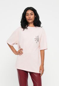 Fekete hajú nő, aki túlságosan nagy, világos rózsaszín T-shirtöt visel virágos mintával és sötétvörös bőrnadrágot, kézzel a csípőjén áll.