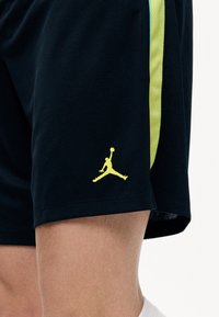 Μαύρα αθλητικά σορτς με κατακόρυφη κίτρινη ρίγα και μικρό κίτρινο λογότυπο Jumpman κοντά στο τελείωμα πάνω από το γόνατο.