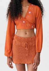 Orange kortärmad topp med långa ärmar har broderade solmotiv i gult och rosa; parat med en matchande virkade mini kjol med snörning.