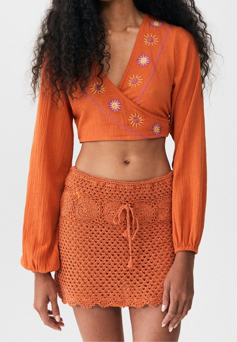 Orange kortärmad topp med långa ärmar har broderade solmotiv i gult och rosa; parat med en matchande virkade mini kjol med snörning.