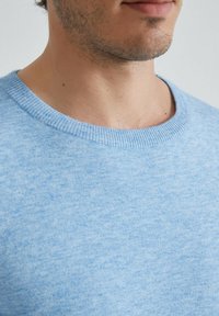 Pull en maille douce, bleu clair, avec un col rond. Le tissu texturé en mélange offre des variations subtiles de couleur et un ajustement confortable.