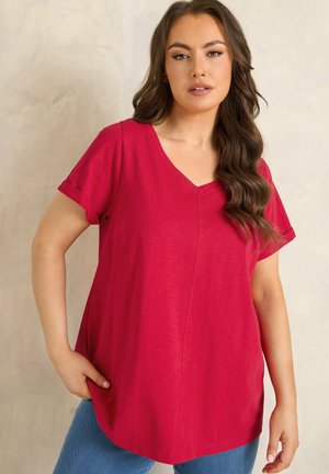 Donna con capelli castani lunghi e mossi, indossa una maglia rossa a maniche corte con scollo a V e jeans blu, in piedi davanti a uno sfondo neutro.