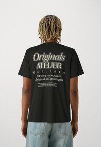 Μαύρη βαμβακερή μπλούζα Τ-shirt με μεγάλη λευκή γραφή στην πλάτη, περιλαμβάνοντας το "Originals ATELIER," με χαλαρή γραμμή και κοντά μανίκια.