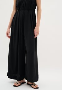 Svart jumpsuit med en avslappnad passform, åtspänd vid midjan med en dragsko. Tillverkad av lättviktigt tyg, med vida ben och minimal textur.
