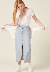 Vaaleansininen farkuhame, jossa on etuaukko, yhdistetty valkoiseen t-paitaan, jota mallin kädet pitävät. Hameessa on korkea vyötärö ja rento suunnittelu.