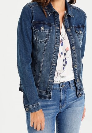 Kobieta w niebieskiej kurtce dżinsowej na białej bluzce w kwiaty i niebieskich dżinsach, ręką swobodnie opuszczoną wzdłuż ciała.