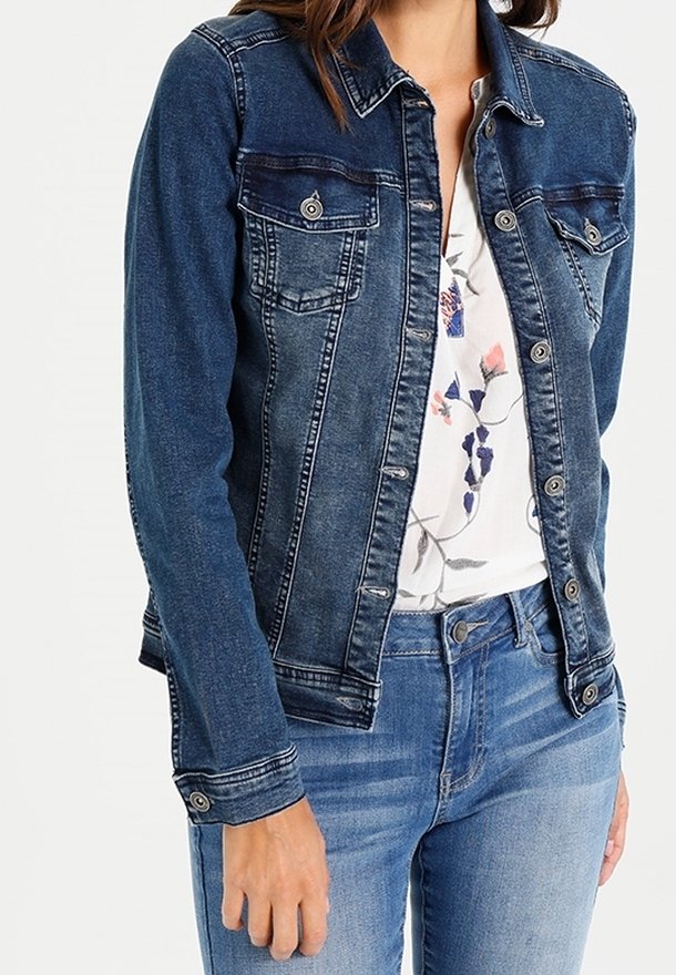 Femme portant une veste en denim bleu sur un chemisier blanc à imprimé floral et un jean bleu, main détendue le long du corps.