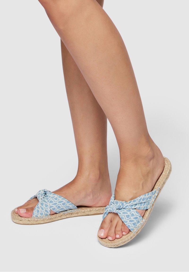 Des pieds portant des sandales à enfiler avec des brides en tissu bleu clair à motifs et des semelles en jute tissé sur un fond uni.