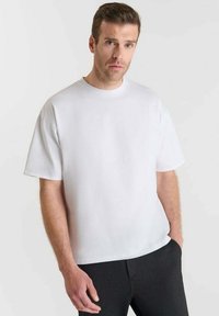 Uomo con capelli corti castani, che indossa una maglietta bianca semplice e oversize e pantaloni neri, in piedi su uno sfondo chiaro.