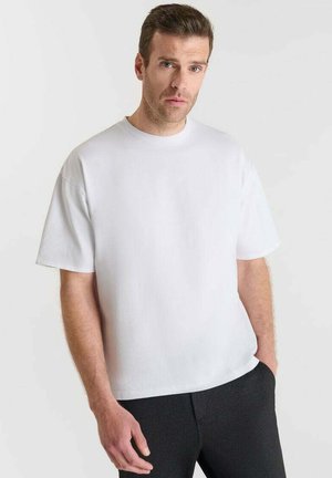 Hombre con cabello corto castaño, vistiendo una camiseta blanca básica de gran tamaño y pantalones negros, de pie contra un fondo claro.