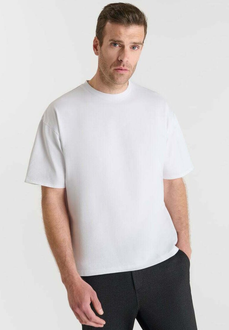 Uomo con capelli corti castani, che indossa una maglietta bianca semplice e oversize e pantaloni neri, in piedi su uno sfondo chiaro.