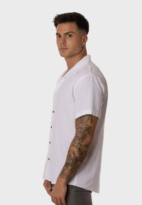 Camisa de lino blanca de manga corta con un diseño de botones, cuello extendido y un ajuste relajado, usada con jeans grises y tatuajes visibles.