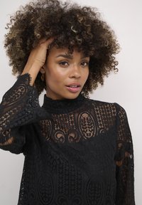 Black lace top with high neckline, intricate patterns, long sleeves, and textured detailing. Model has curly hair, highlighted strands.