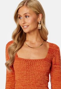 Maglione a coste di colore arancione con scollatura quadrata. Indossa una collana in tono oro e grandi orecchini texture. Capelli in onde morbide.