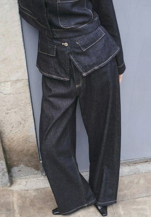 Persona vistiendo pantalones anchos de mezclilla oscura y chaqueta a juego con bolsillos con solapa, de pie frente a un fondo de piedra y metal.