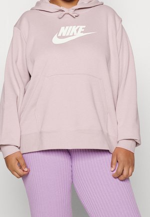 Person iført en lyserød Nike hættetrøje med logo og lavendel ribbukser, stående mod en ensfarvet lys baggrund.