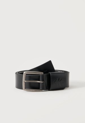 Ceinture en cuir noire avec une boucle en métal argenté et un logo Calvin Klein embossé près de la boucle, enroulée sur un fond blanc.