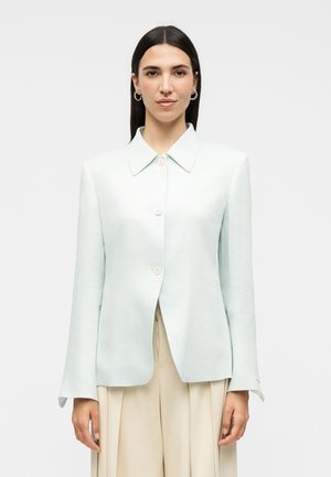 Γυναίκα με ίσια σκούρα μαλλιά φοράει ανοιχτό μπλε σακάκι με κουμπιά και μπεζ πλισέ παντελόνες, κοιτάζοντας μπροστά με ουδέτερη έκφραση.