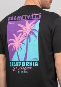 T-shirt noir avec un design graphique comprenant des palmiers roses, un fond bleu, et du texte rétro : "Palm Beach" et "California."