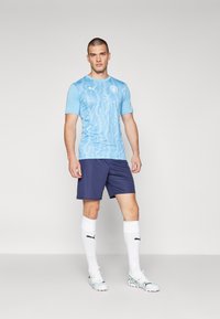 Maillot de football bleu clair avec un design à motifs, shorts navy, chaussettes blanches et crampons argentés, arborant des accents du logo noir de Puma.