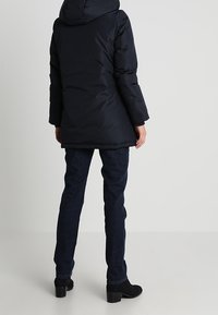 Lange zwarte donsjack met een capuchon, voorzien van een gladde, glanzende afwerking. Gecombineerd met donkerblauwe jeans en zwarte enkellaarsjes.