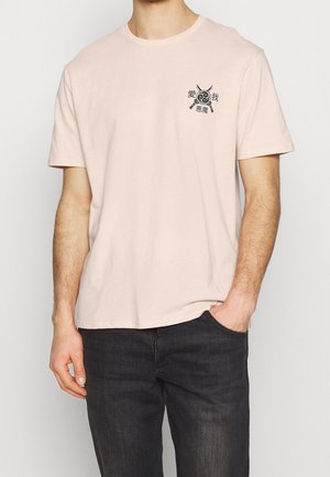 Beige T-shirt met korte mouwen met een afbeelding van gekruiste zwaarden en een cirkelvormig embleem op de linkerborst, gedragen door een persoon met de rechterhand in de zak van een zwarte spijkerbroek.