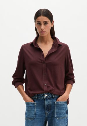 Vrouw met donker haar die een bordeauxrode blouse met knopen en een blauwe spijkerbroek draagt, met handen in de voorzakken tegen een effen achtergrond.