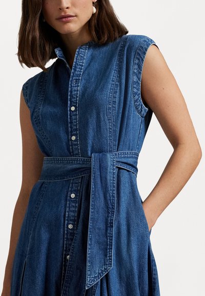 Denim kleita ar īsām piedurknēm, pogu aizdari un sasietu jostu. Iekļauj strukturētu apkakli un pleitu svārkus tumši zilā krāsā.