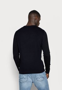 Suéter negro de punto con mangas largas, cuello redondo, puños y dobladillo de canalé, combinado con jeans desgastados de un azul claro, visto desde la parte de atrás.