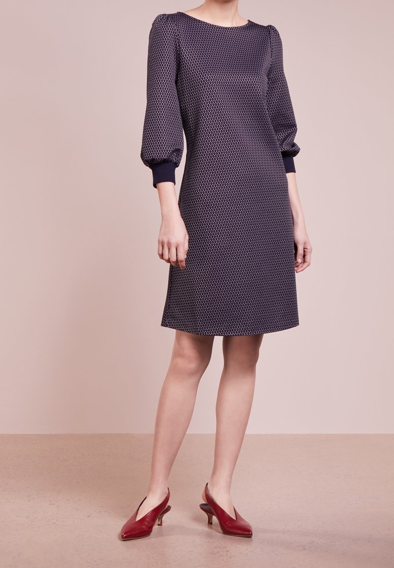 Femme portant une robe bleu marine mi-longue avec de petits pois blancs et des manches bouffantes trois-quarts, associée à des chaussures rouges à talons bas.