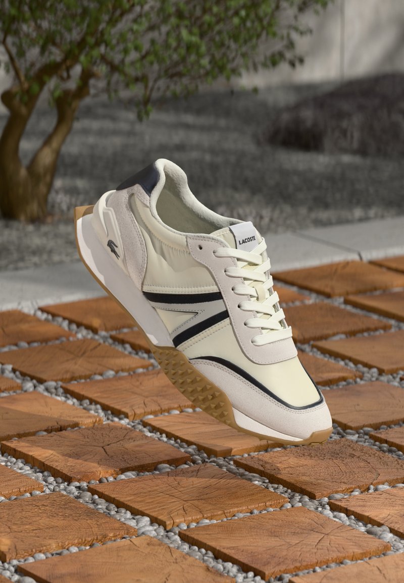 Fehér és bézs Lacoste sportcipő fekete díszítéssel, amely fa négyzet alakú csempék és apró szürke kövek felett lebeg a szabadban.