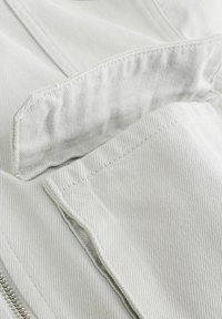 Prim-plan cu material din denim alb, evidențiind cusăturile detaliate și clapeta buzunarului parțial deschisă deasupra unui fermoar vizibil.