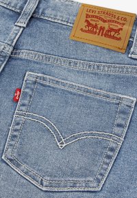 Ανοιχτό μπλε τζιν παντελόνι με πίσω τσέπη που διαθέτει χαρακτηριστικό μοτίβο ραφής, εμβληματική κόκκινη ετικέτα και δερμάτινο ταμπελάκι με το λογότυπο της μάρκας.