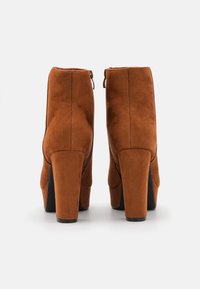 Botas de tornozelo em camurça castanha com um alto salto empilhado, apresentando um fecho de zipper na parte traseira e um design elegante e afunilado.
