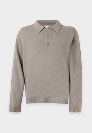 Jeune homme debout portant un pull beige à col boutonné, un pantalon ample vert clair et des chaussures marron foncé sur un fond blanc uni.
