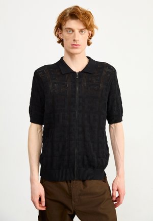 Gilet zippé noir à manches courtes avec un motif tricoté texturé et un col à côtes. Présente un design géométrique complexe sur l'ensemble.