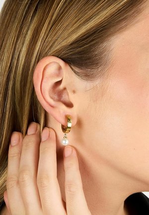 Nærbillede af en hånd, der rører ved en gylden ørering med en hængende hvid perle på en kvindes øre og lyst brunt hår.