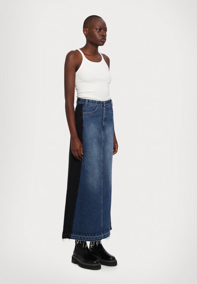 Denim maxi szoknya kék elülső résszel és fekete oldalpannelsel, bolyhos szegéllyel és klasszikus öt zsebes kialakítással. Fehér ujjatlan toppal viselve.