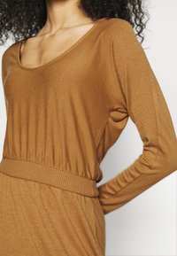 Haut brun à manches longues en tissu doux avec un décolleté échancré, une taille côtelée et une coupe drapée. La finition texturée ajoute une profondeur subtile.