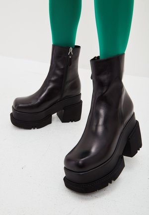 Schwarze klobige Plateau-Stiefeletten mit dicken Blockabsätzen und seitlichen Reißverschlüssen, getragen mit grünen Strumpfhosen auf weißem Boden.