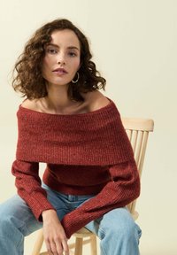 Maglione rosso a spalle scoperte, a coste, con maniche lunghe, abbinato a jeans blu chiaro, seduto su una sedia di legno contro uno sfondo beige.