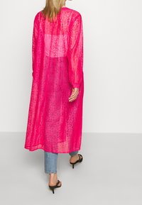 Cardigan lungo e trasparente di colore rosa, con un motivo testurizzato, dotato di maniche lunghe e una vestibilità comoda, abbinato a jeans blu e tacchi neri.