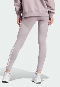 Ljust lila leggings i stretchmaterial med en vit logotyp längs vänster ben, ihopparat med vita sportskor.