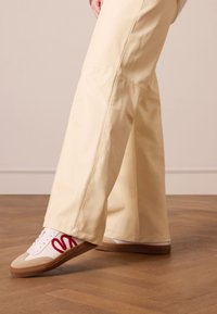 Cream-colored wide-leg pants paired with white sneakers featuring red detailing and beige accents. Wooden floor background.