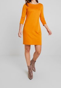 Orange fitted dress with three-quarter sleeves, smooth texture, knee-length, paired with beige snake-print ankle boots.