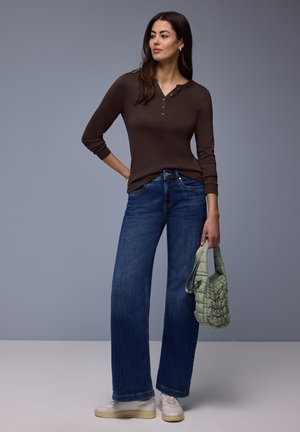 Kobieta ubrana w brązową bluzkę henley z długim rękawem, szerokie niebieskie dżinsy, białe trampki, trzymająca małą zieloną pikowaną torebkę, stojąca na tle szarego tła.