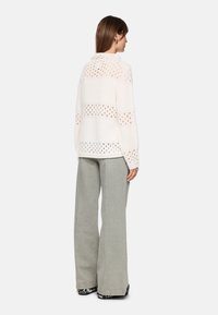 Cremfarbener Strickpullover mit offenen Mustern, langen Ärmeln und lockerer Passform, kombiniert mit hellgrauen weiten Hosen und Schuhen im Leopardenmuster.
